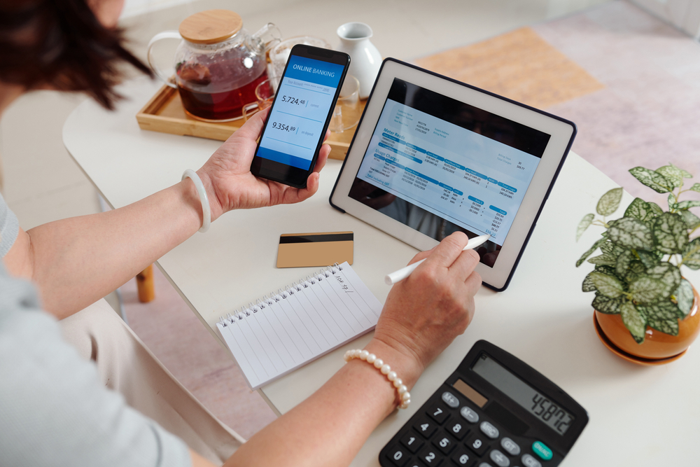Eine Person am Schreibtisch hält ein Handy mit Online Banking in der Hand und ein Tablet auf dem Tisch