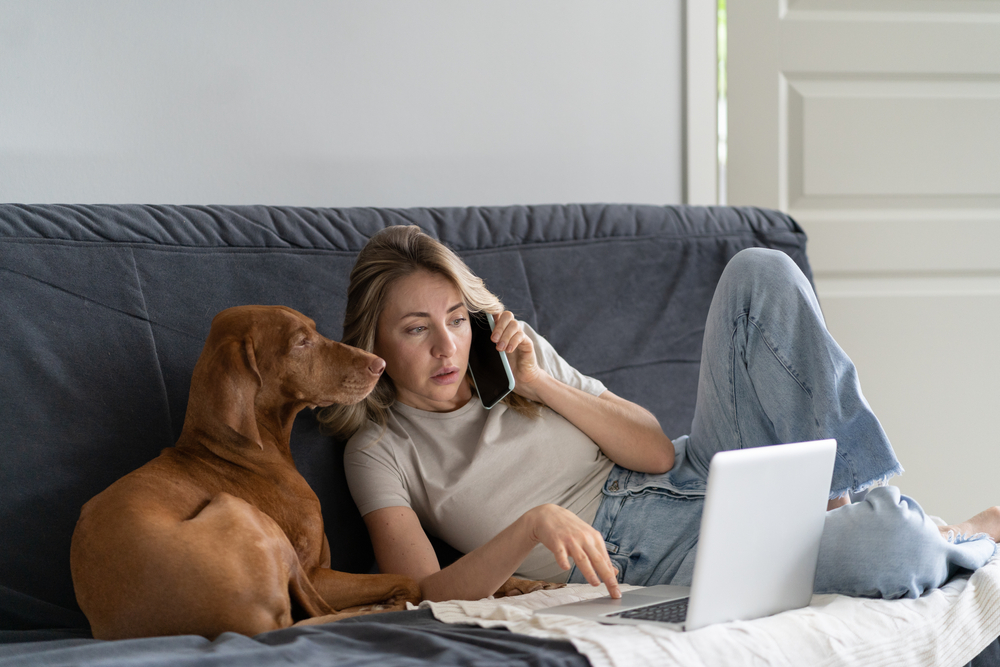 Telefonierende Frau mit Hund und LapTop am Sofa
